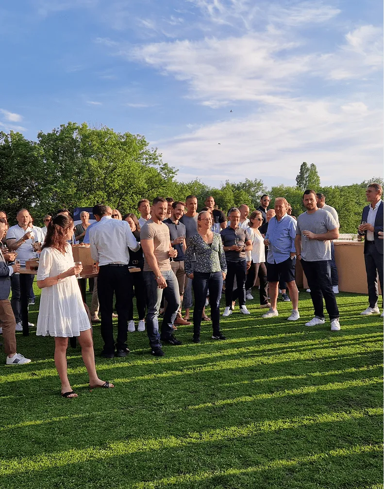 Group of people socializing outdoors on green grass under a blue sky with scattered clouds.