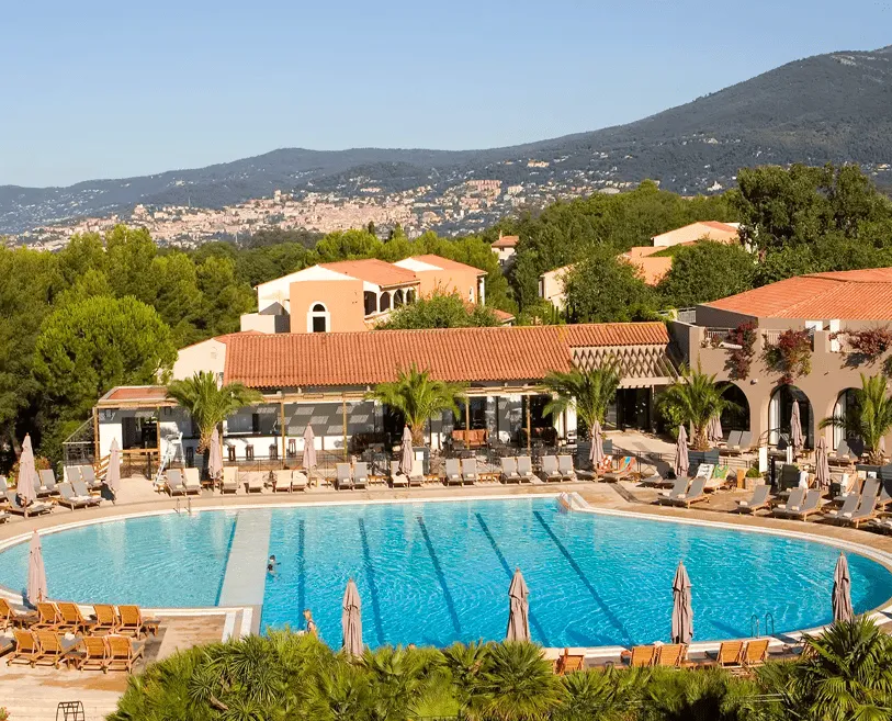 Outdoor swimming pool surrounded by lounge chairs and umbrellas with terracotta-roofed buildings and green trees, set against a mountainous backdrop.
