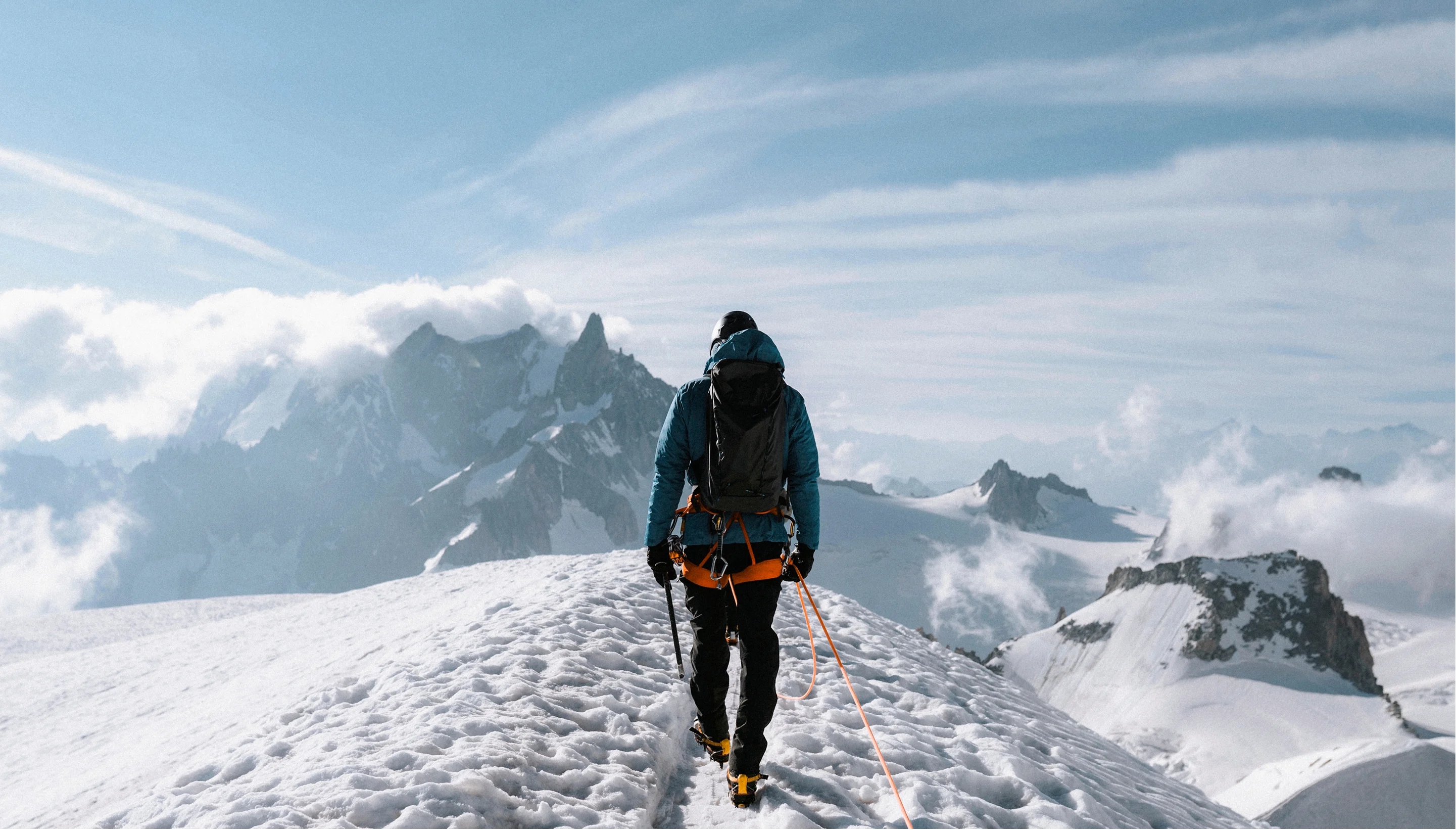 Mountaineer in blue jacket hiking on snow-covered ridge with mountain peaks and clouds in background.