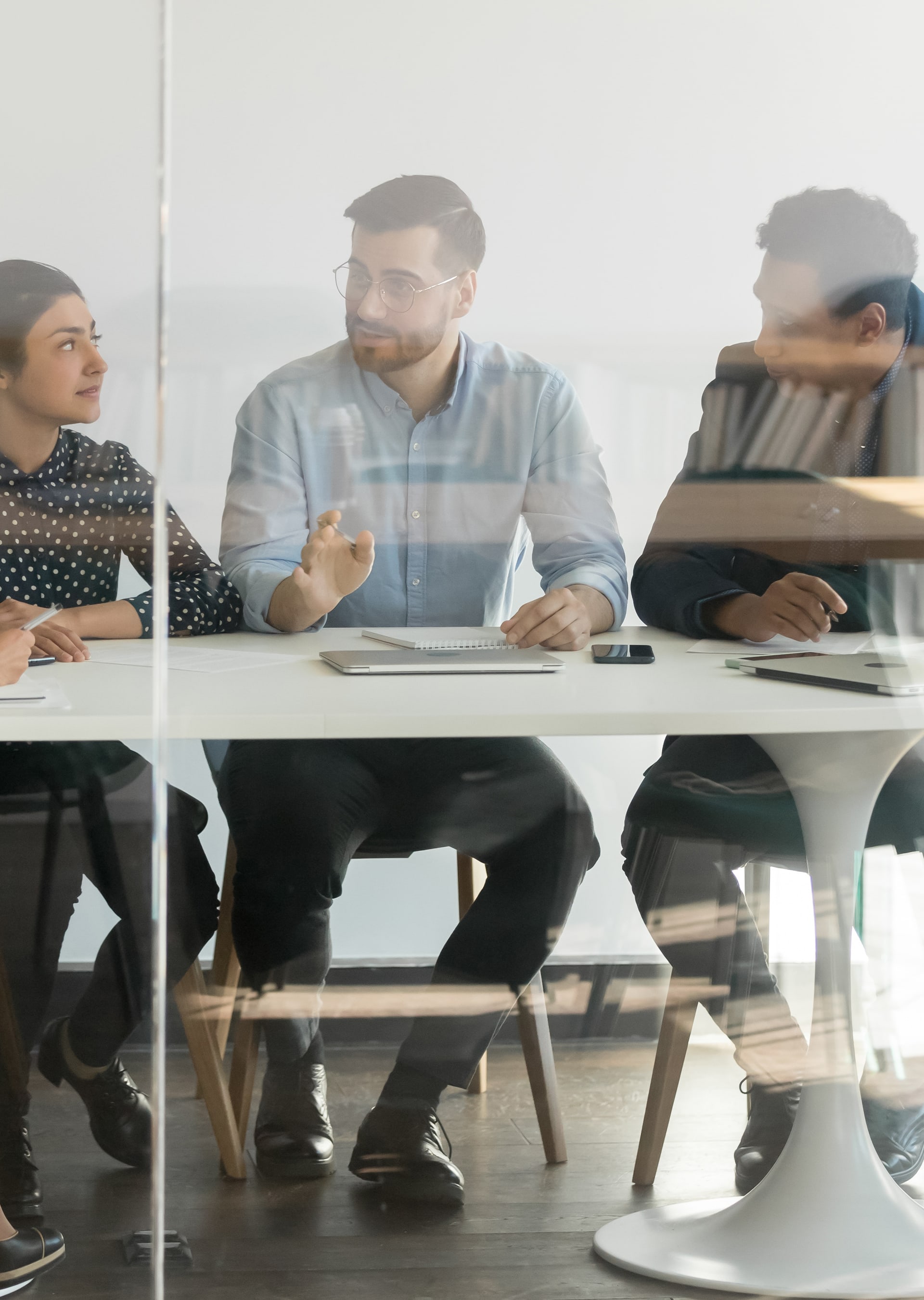 Three colleagues sitting at a white table engaged in discussion behind a glass wall in an office.