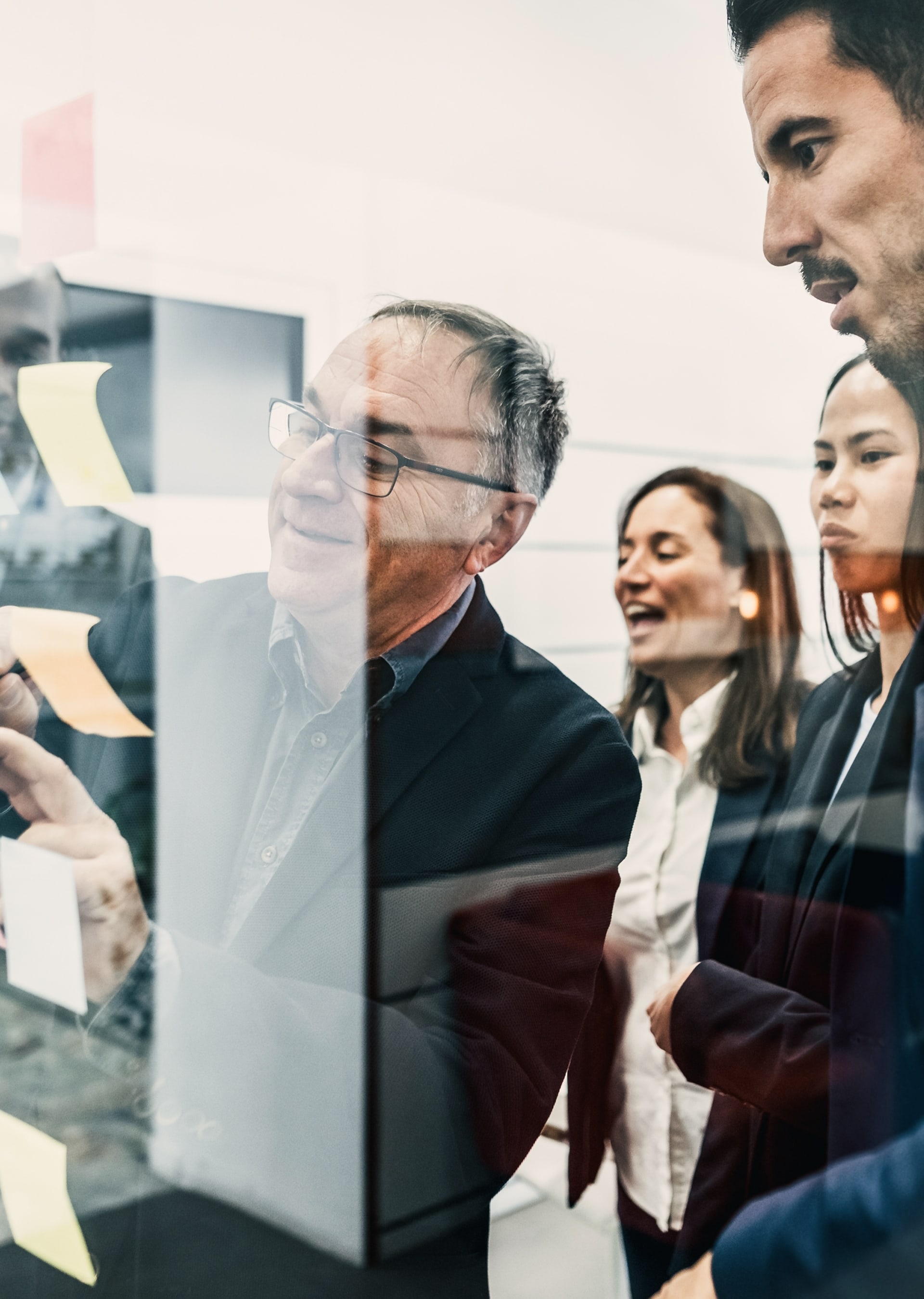 Group of four diverse business professionals collaborating near a glass board with sticky notes.