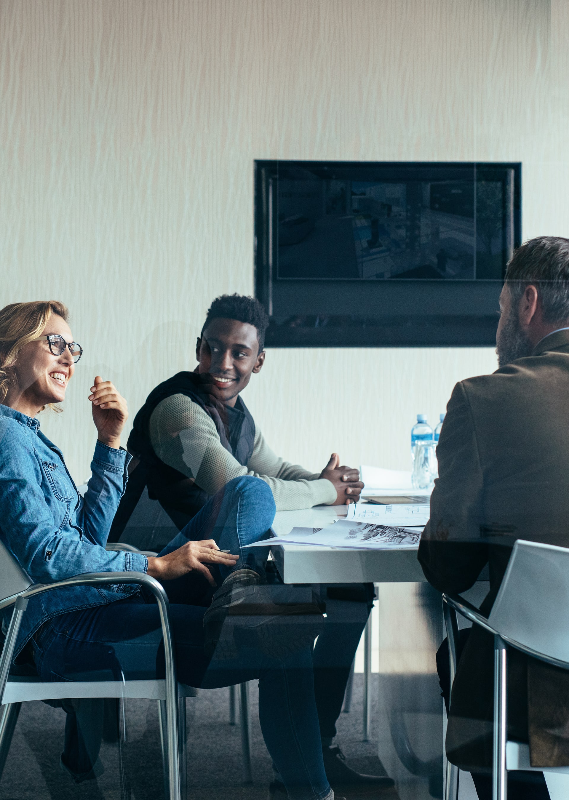 Three colleagues sitting around a table in a meeting room, discussing work with papers and water bottles on the table.