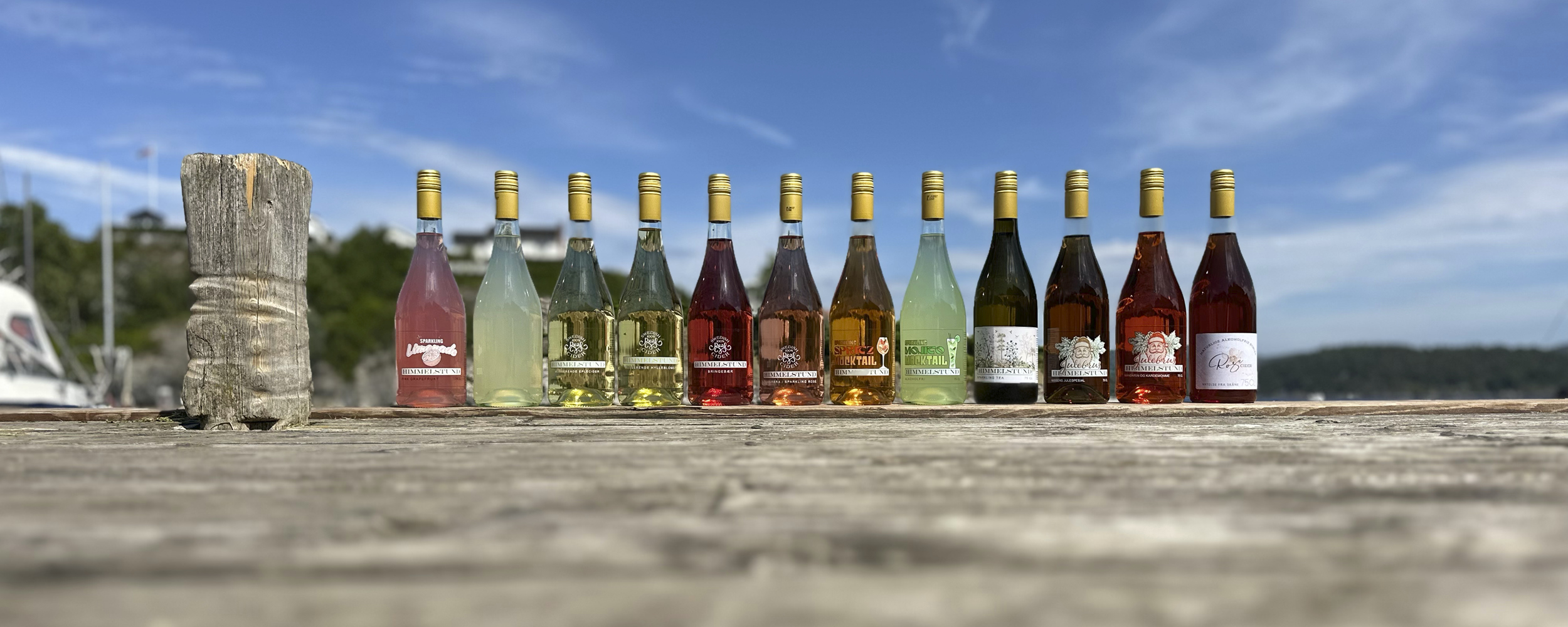 En rekke fargerike flasker med alkoholholdige drikker står på et trebord utendørs under en blå himmel.