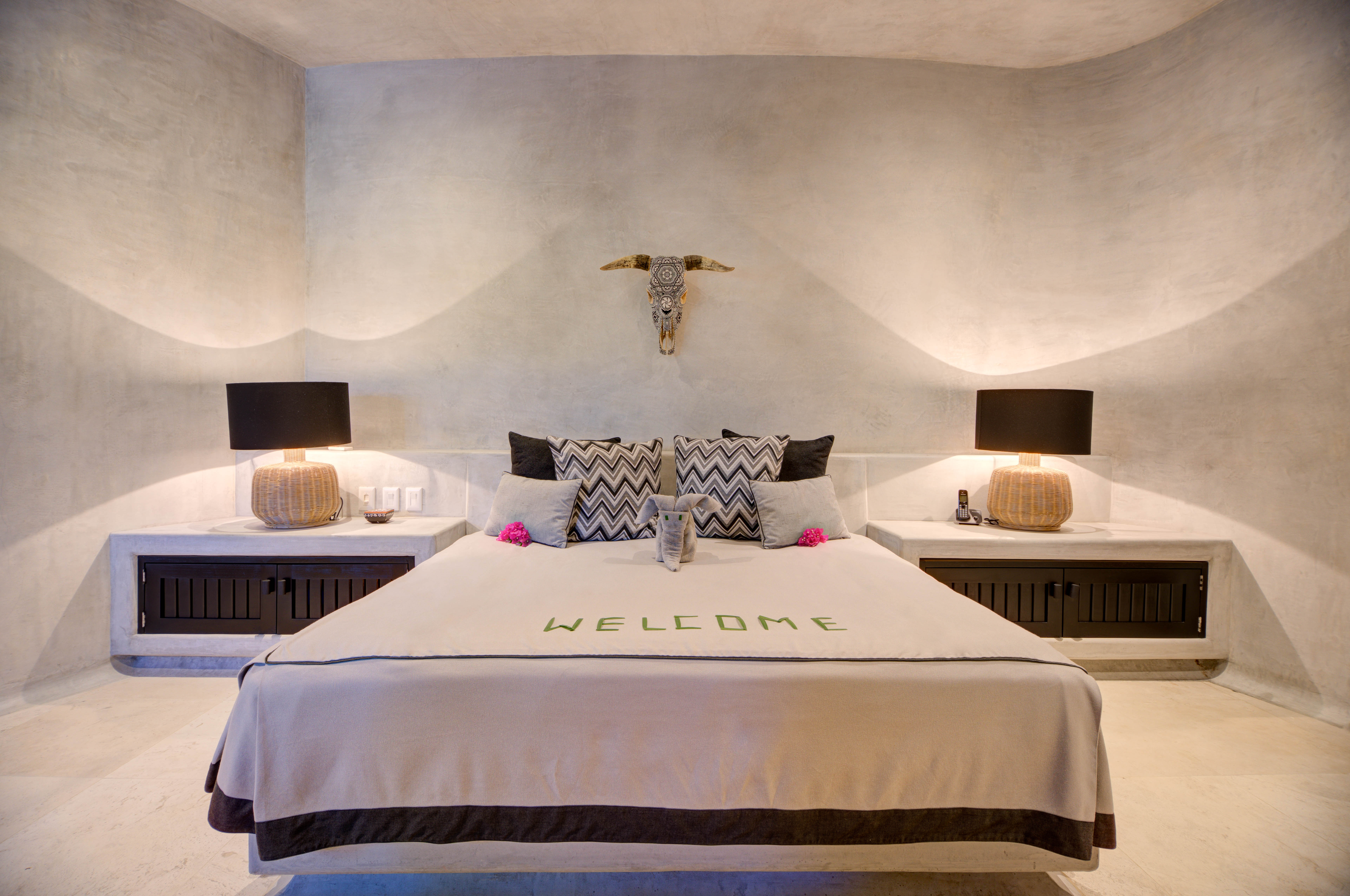 Cozy bedroom with red-accented pillows, large mirrors on walls, and sliding shutters opening to a lit pool and ocean view at dusk.