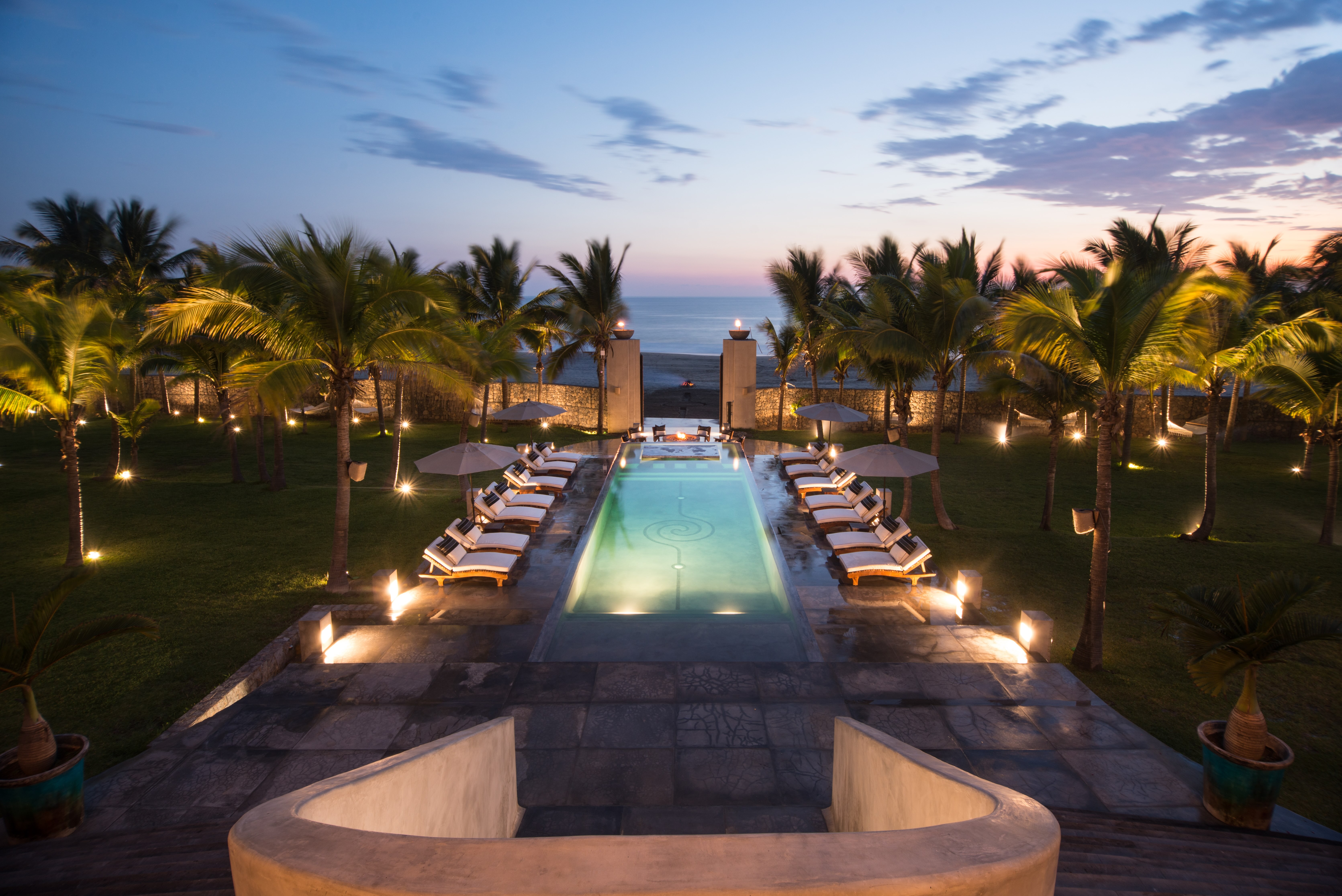 Infinity pool with lounge chairs and umbrellas on both sides, palm trees in the background, and a person meditating at the edge facing the ocean.