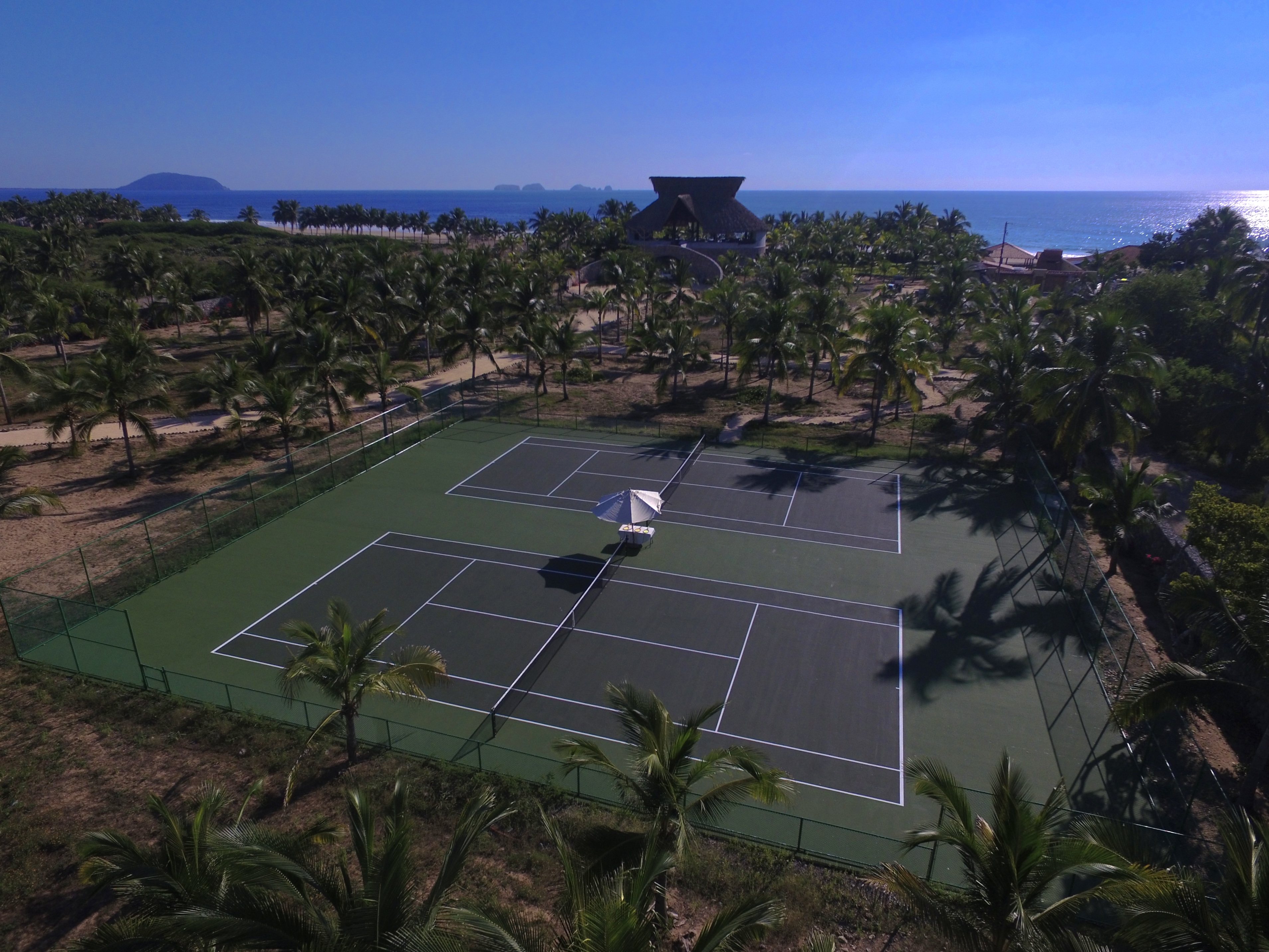 Aerial view of two adjacent fenced tennis courts surrounded by palm trees near a sandy beach and ocean.