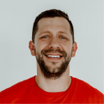 Smiling man with short dark hair and beard wearing a red shirt against a plain background.
