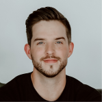 Portrait of a young man with short dark hair, a beard, and a mustache, wearing a black shirt against a light gray background.