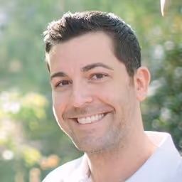 Smiling man with short dark hair and light stubble, wearing a white shirt, outdoors with blurred greenery background.