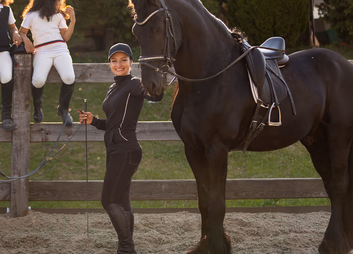 Smiling woman in equestrian gear holding reins and touching the head of a black horse in an outdoor paddock.