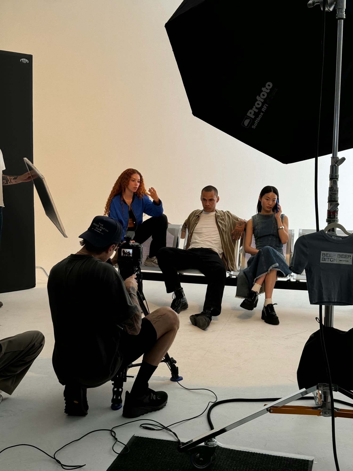 Camera operator crouched behind a tripod filming three models on metal bench seats in a studio lit by a large octabox