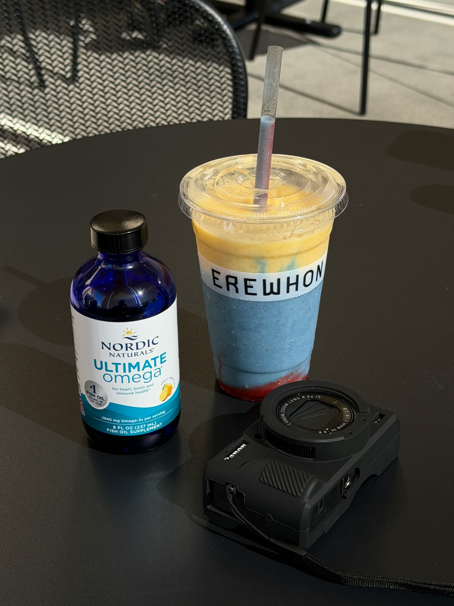 Bottle of fish oil supplement beside a layered blue and yellow smoothie and a compact camera on a black cafe table
