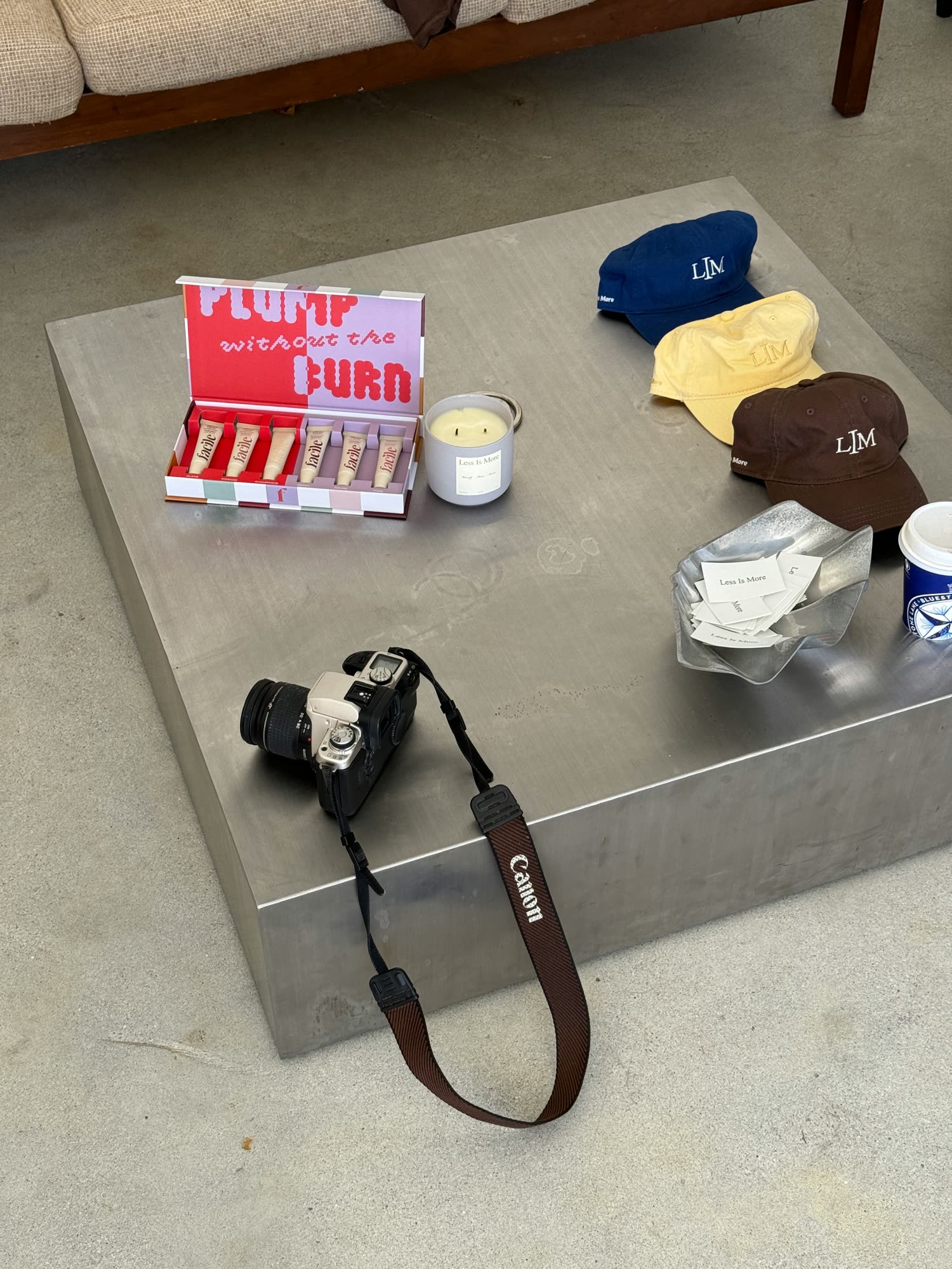 Open gift box of six lip tubes beside a candle, three monogrammed caps, a bowl of cards and a film camera on a steel plinth