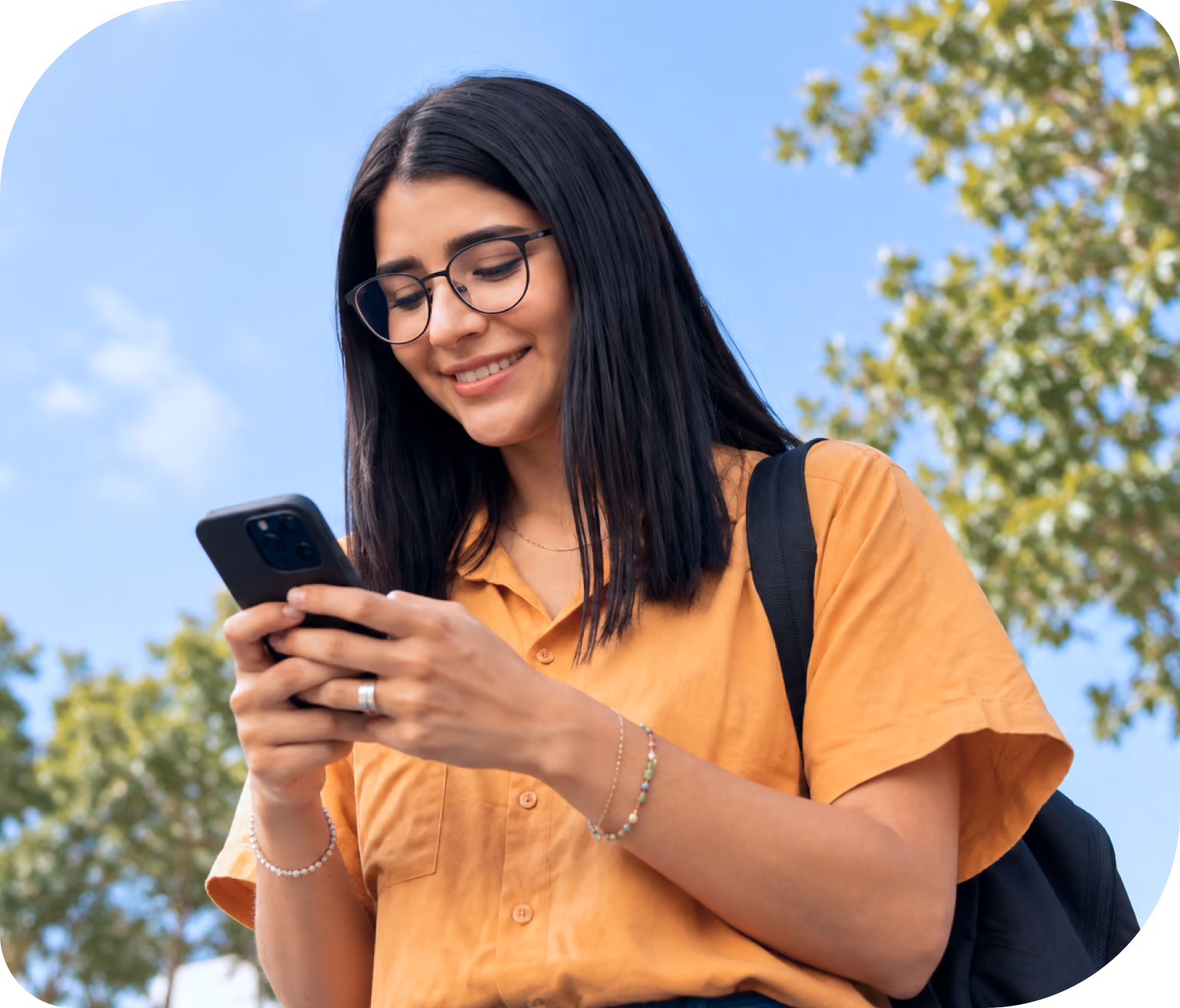 Joven sonriente con gafas y camisa naranja usando un teléfono móvil al aire libre con árboles y cielo azul de fondo.