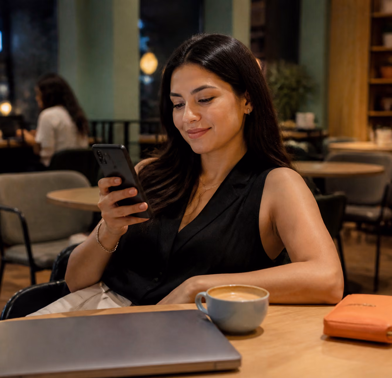 Mujer sentada en cafetería mirando su teléfono con café, portátil cerrado y cartera naranja sobre la mesa.