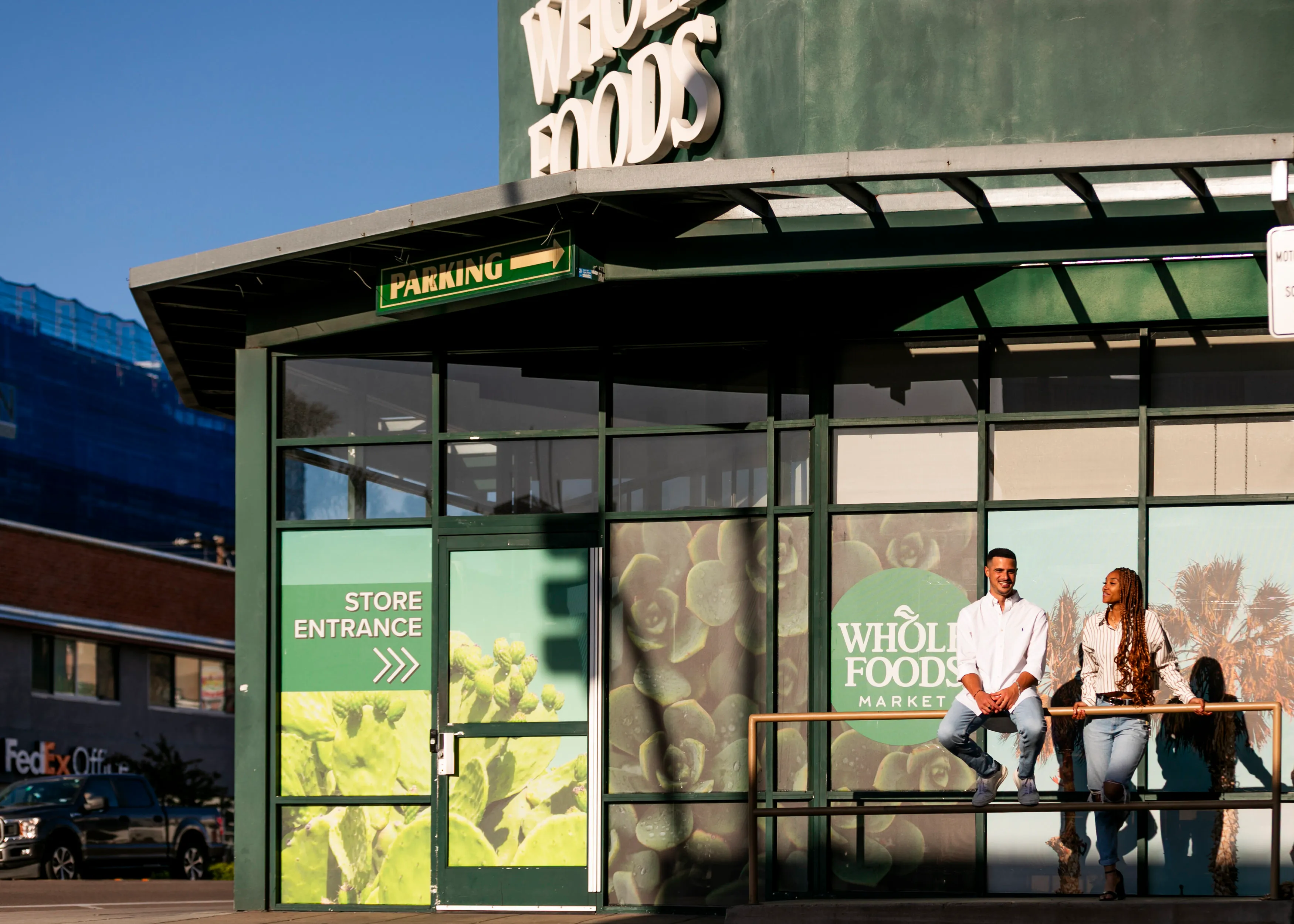 Two people sitting and standing by the railing outside a Whole Foods Market entrance with signage.