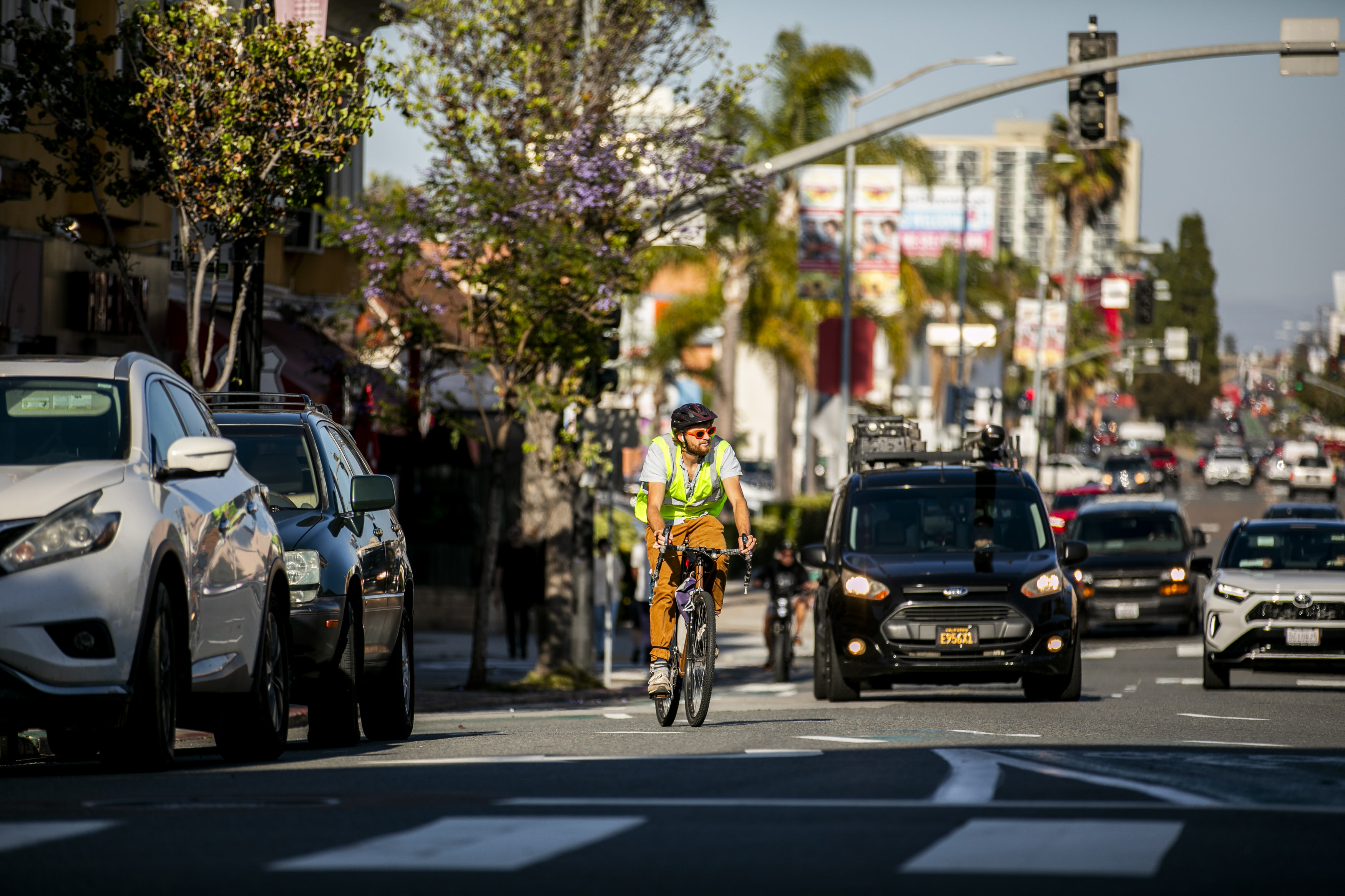 Cyclist wearing a helmet, sunglasses, and a reflective vest riding on a city street with parked cars and traffic in the background.