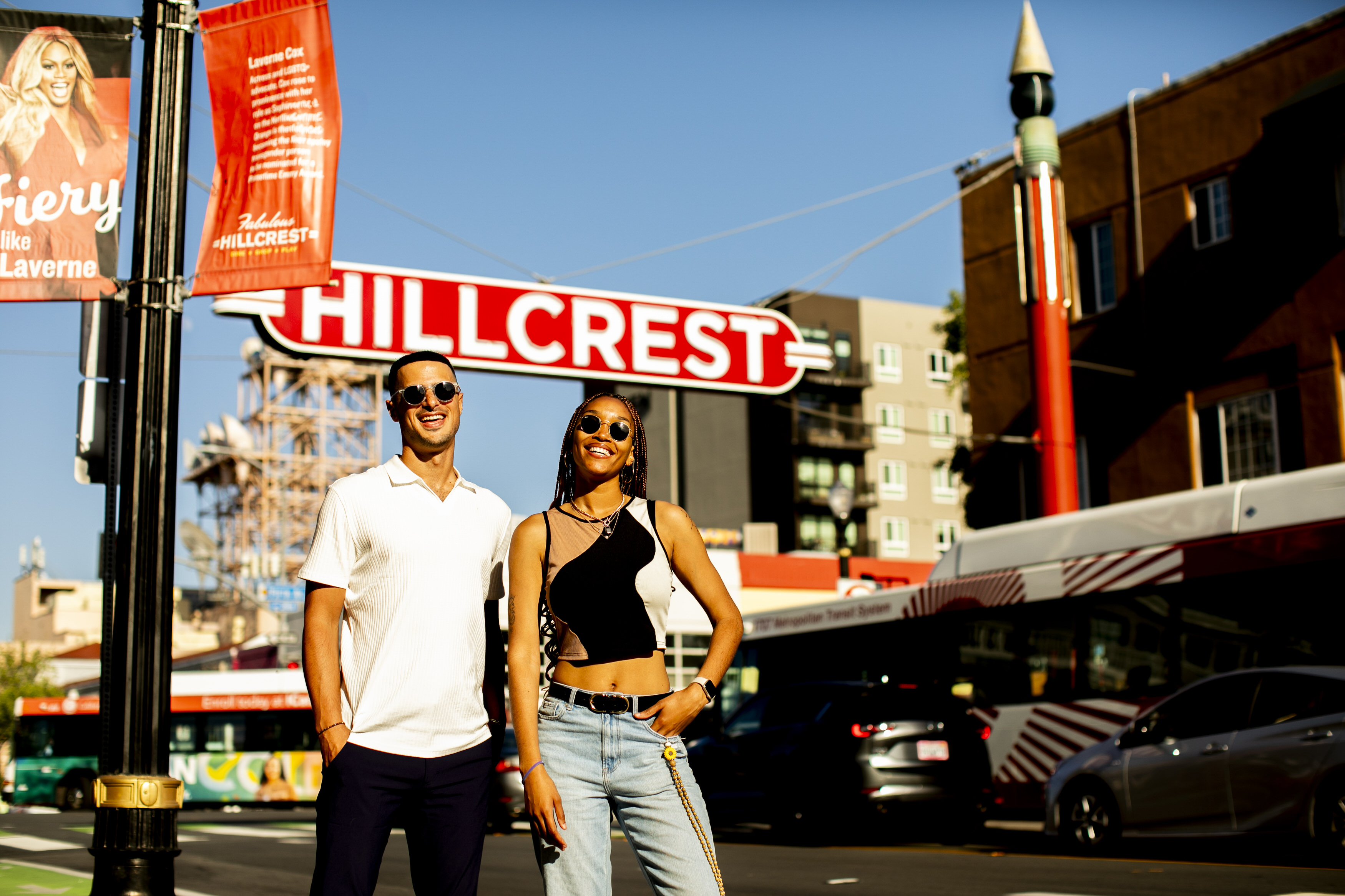 Two smiling young adults wearing sunglasses standing in front of a red Hillcrest sign on a sunny urban street.