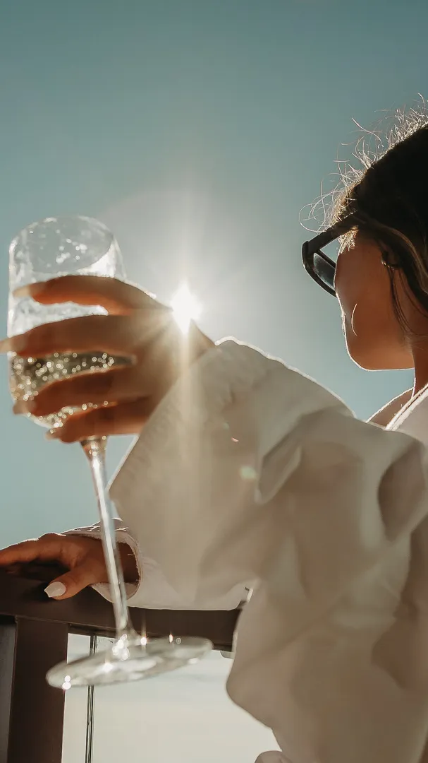 Person in sunglasses and white clothing holding a glass of sparkling wine against a bright blue sky with the sun shining through the glass.