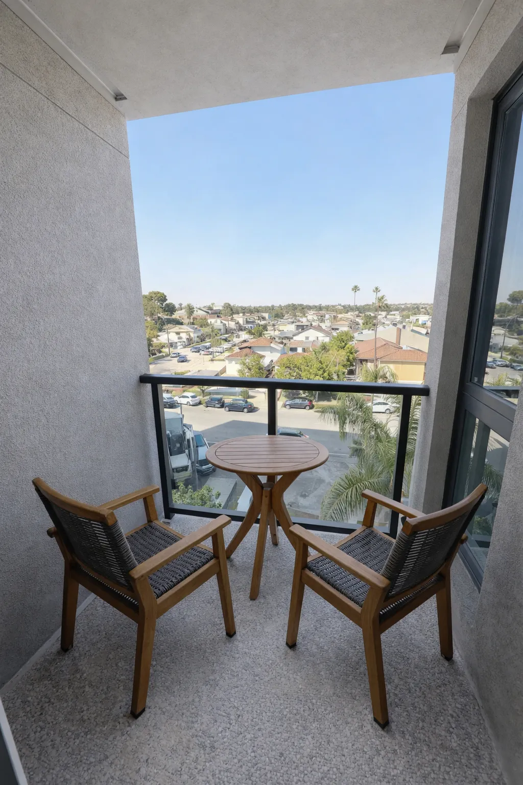 Small balcony with two wooden chairs and a round wooden table overlooking a neighborhood with houses and cars.