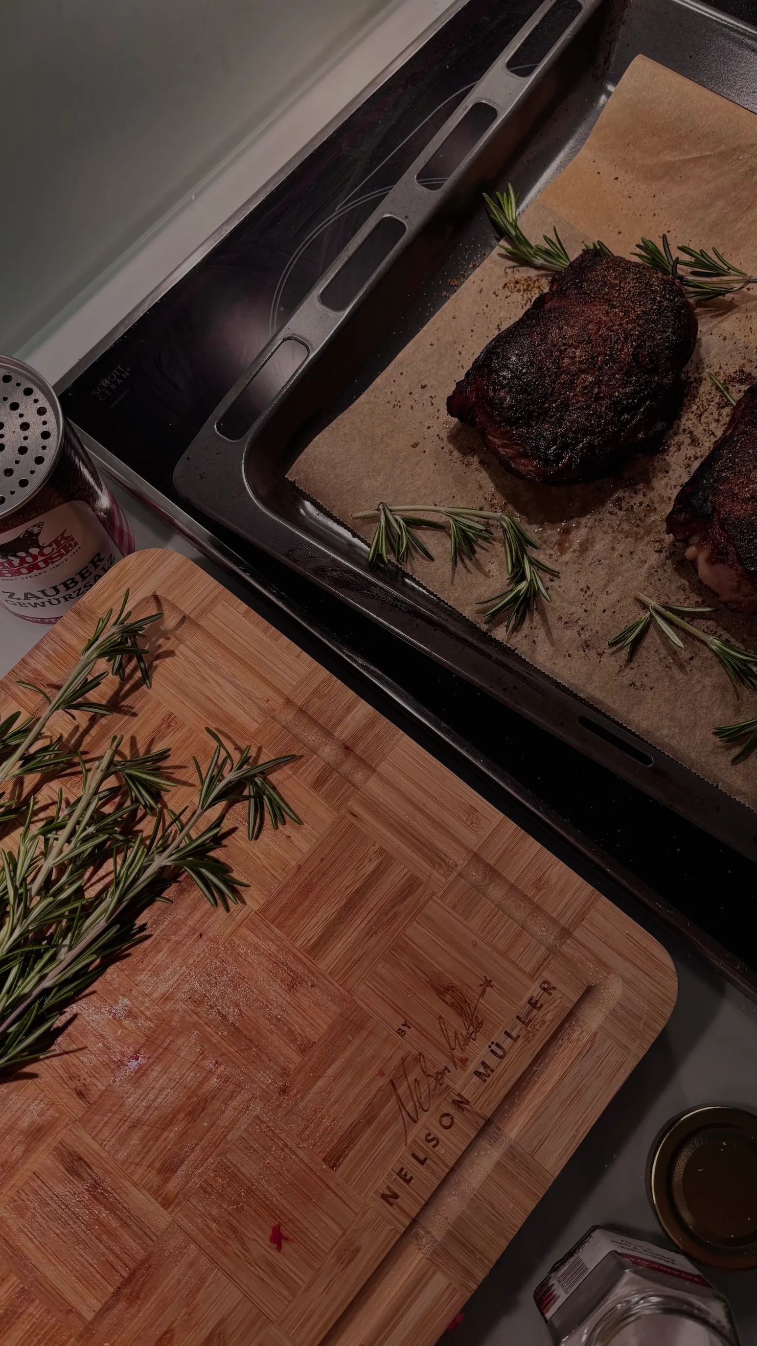 Two cooked steaks with rosemary on parchment in a baking tray next to a wooden cutting board with fresh rosemary and a spice container.