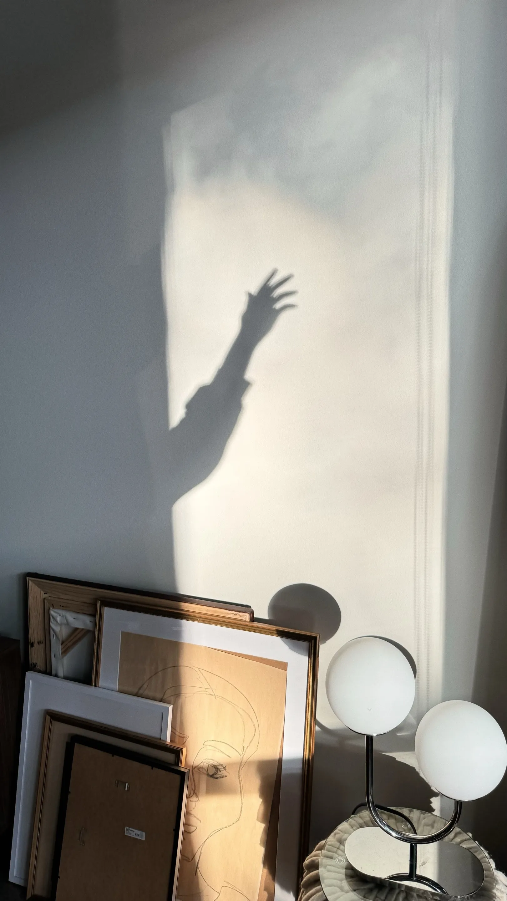Shadow of an arm and hand raised on a wall above framed artwork and a modern table lamp with two round white bulbs.