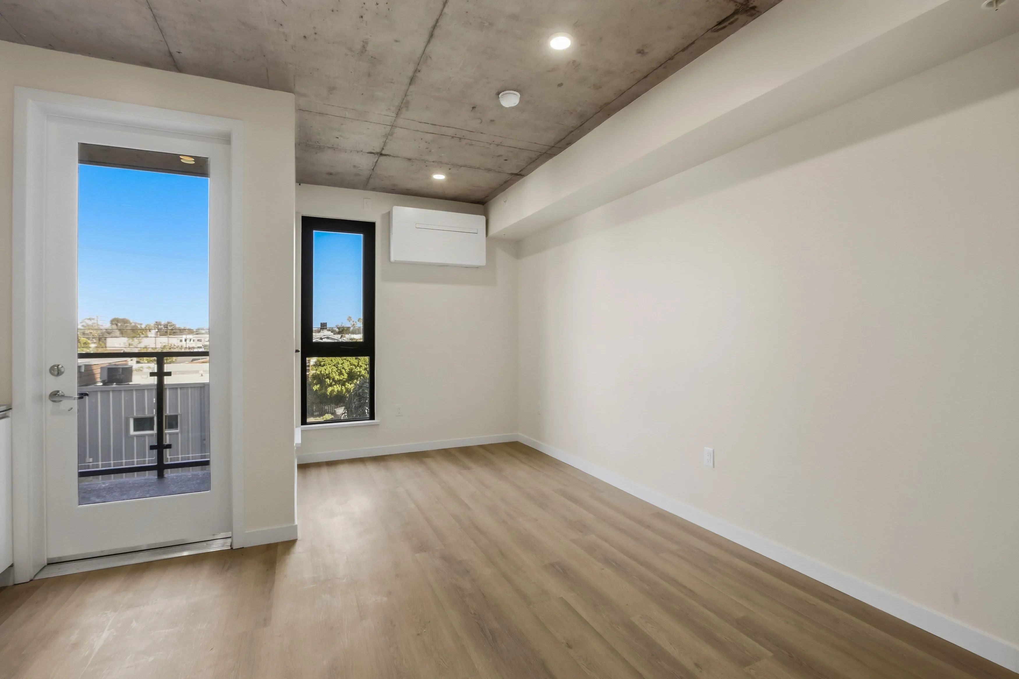 Empty modern apartment room with light wood flooring, off-white walls, exposed concrete ceiling, a large window, and a glass door leading to a balcony.