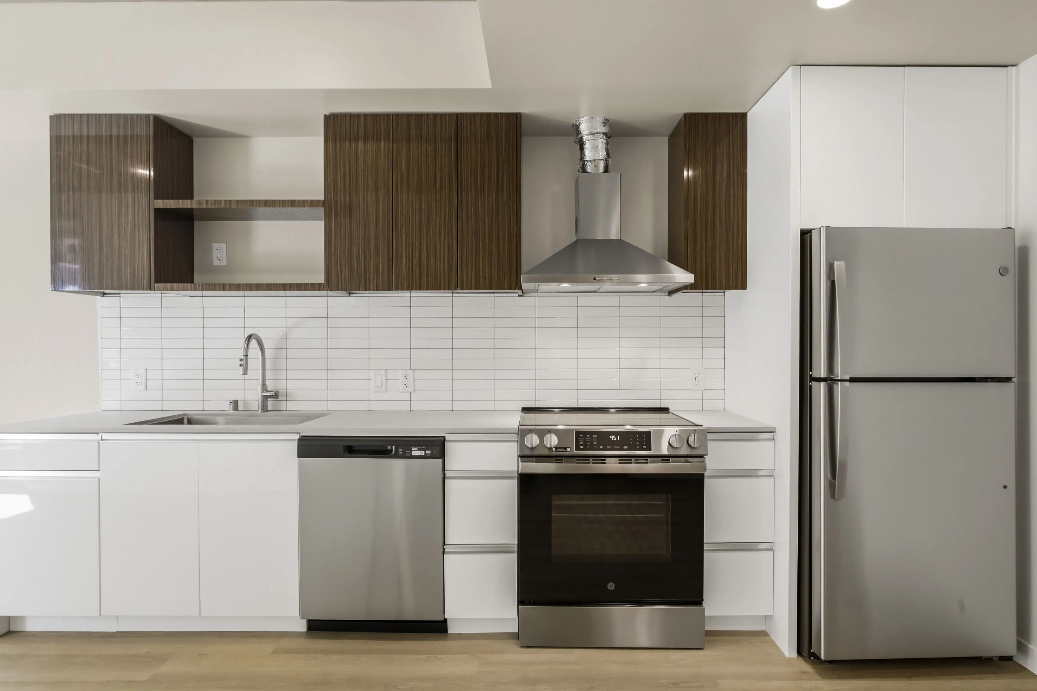 Modern kitchen with white lower cabinets, wood-textured upper cabinets, stainless steel appliances including a refrigerator, dishwasher, oven, and a range hood.
