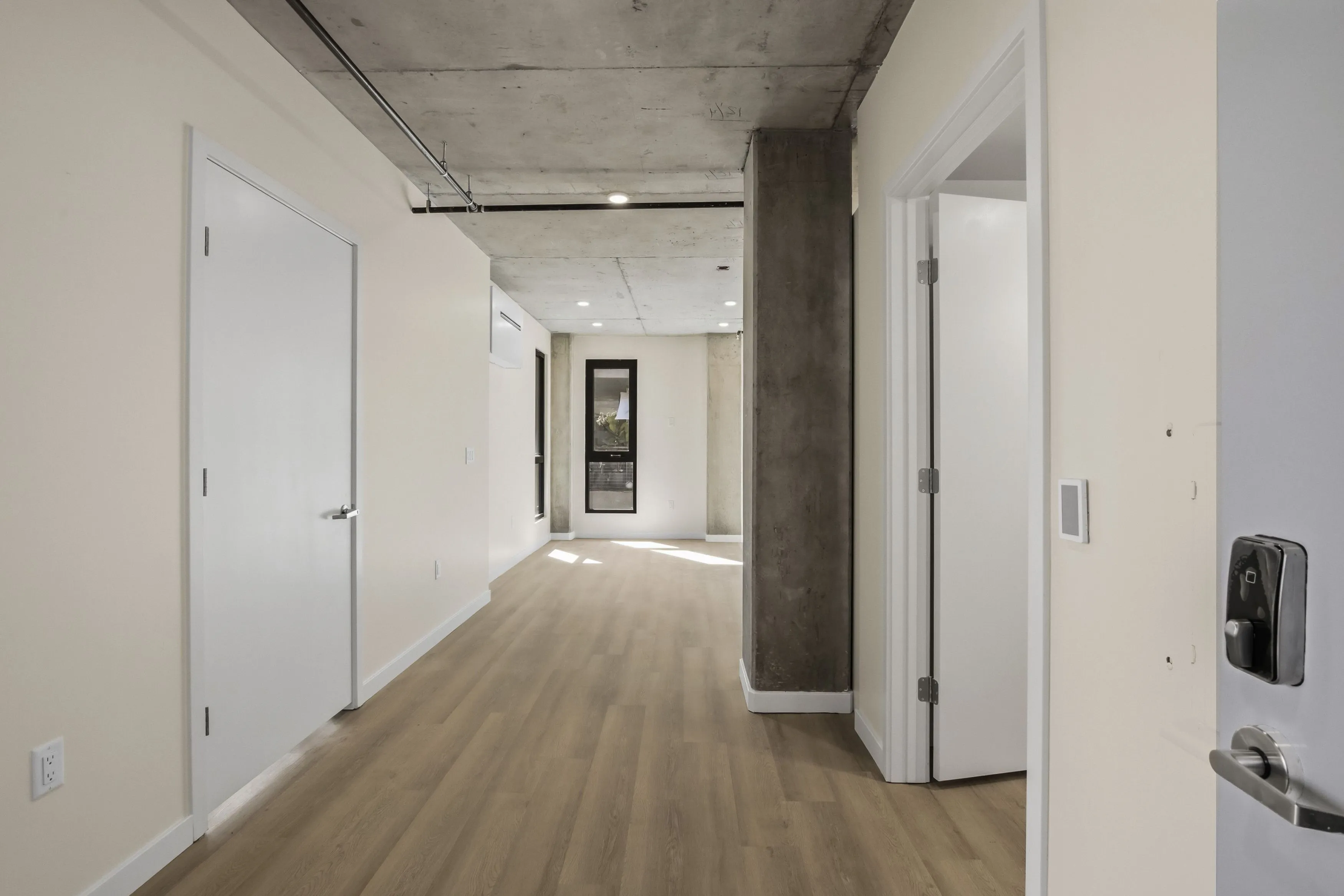 Empty modern apartment hallway with light wood flooring, white doors, and exposed concrete ceiling and column.