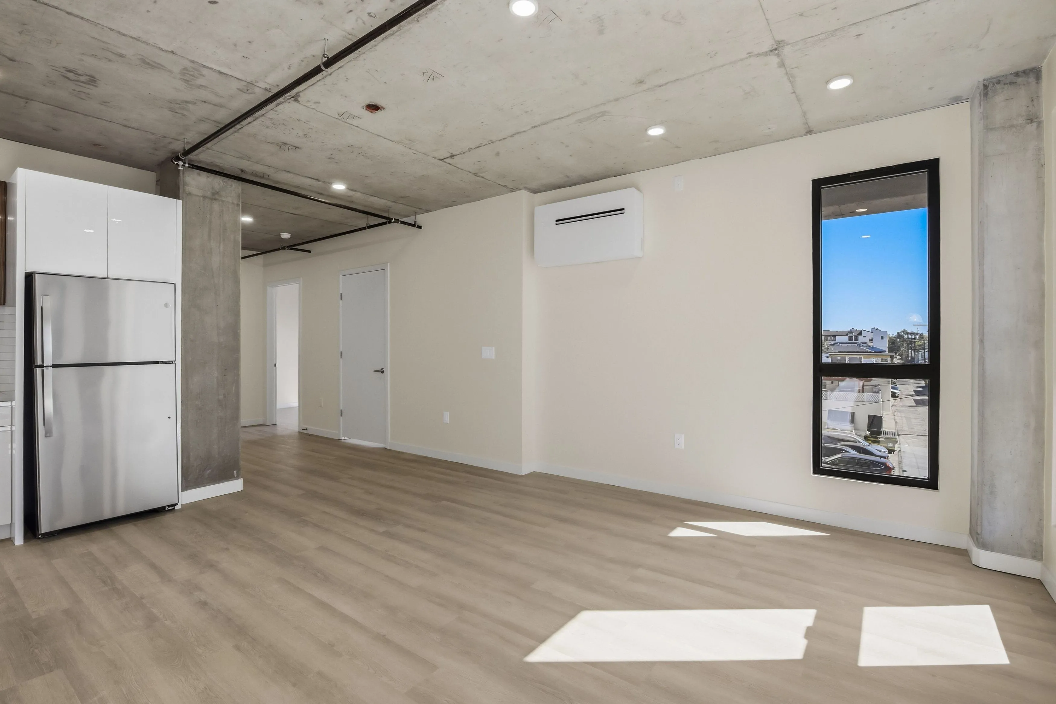 Modern empty apartment living area with light wood flooring, concrete ceiling, stainless steel refrigerator, and a tall narrow window showing a city view.