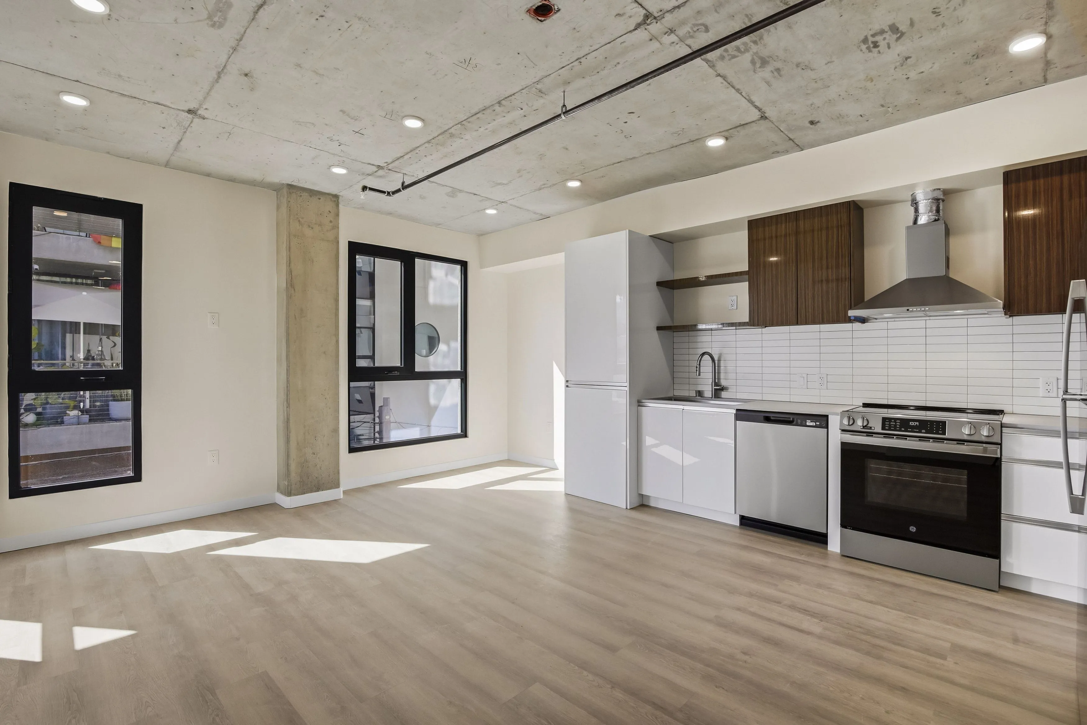 Modern kitchen with light wood flooring, white cabinets, stainless steel appliances, and two large black-framed windows; exposed concrete ceiling with recessed lighting.