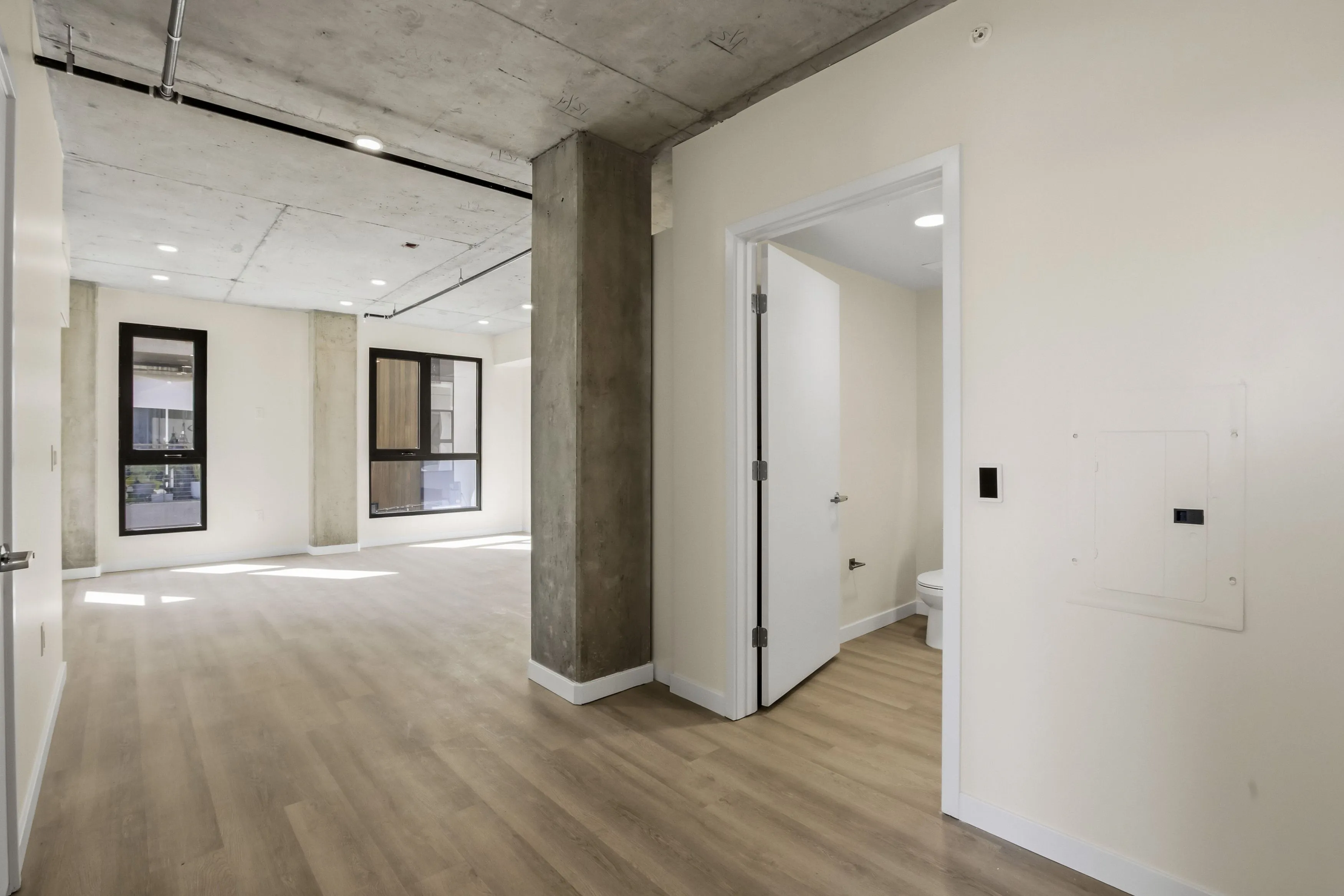 Modern empty apartment interior with light wood flooring, concrete ceiling, large windows, and an open door to a bathroom.