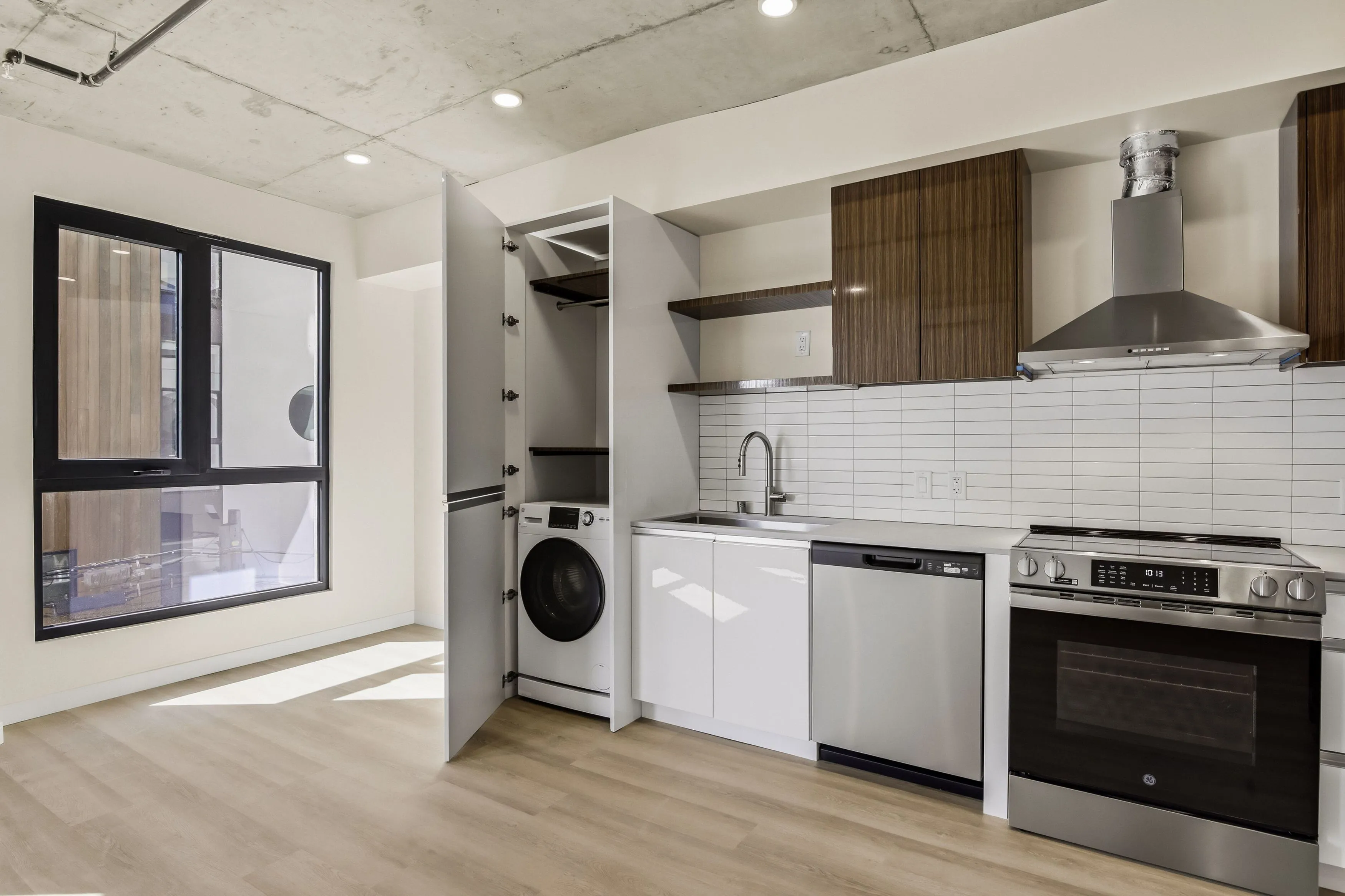 Modern kitchen with light wood flooring, a large window, white cabinets, stainless steel appliances, and a washing machine inside an open cabinet.