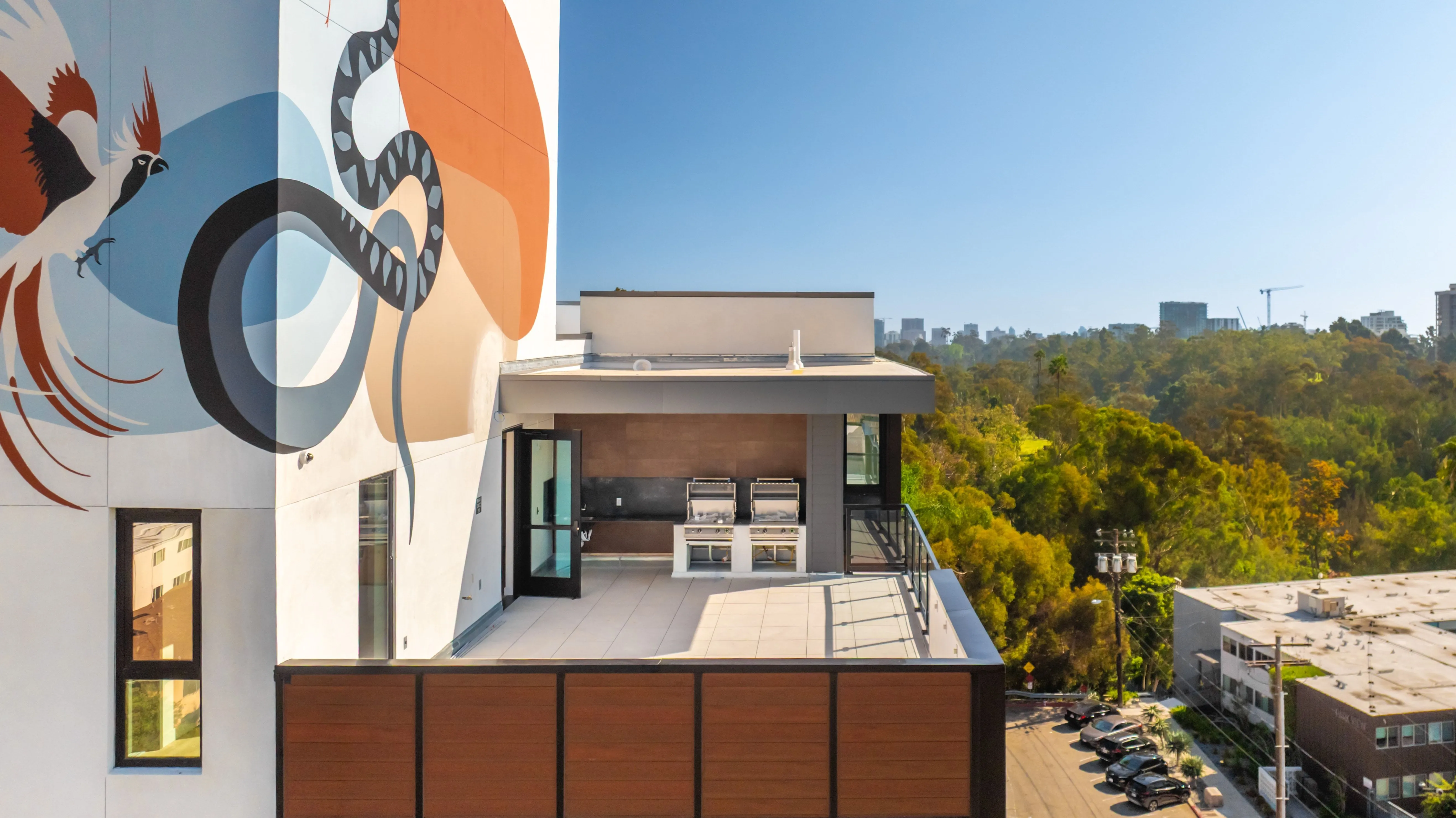 Modern rooftop terrace with two stainless steel grills, large tiled floor, and wall mural featuring a bird and abstract shapes, overlooking a green park and cityscape.