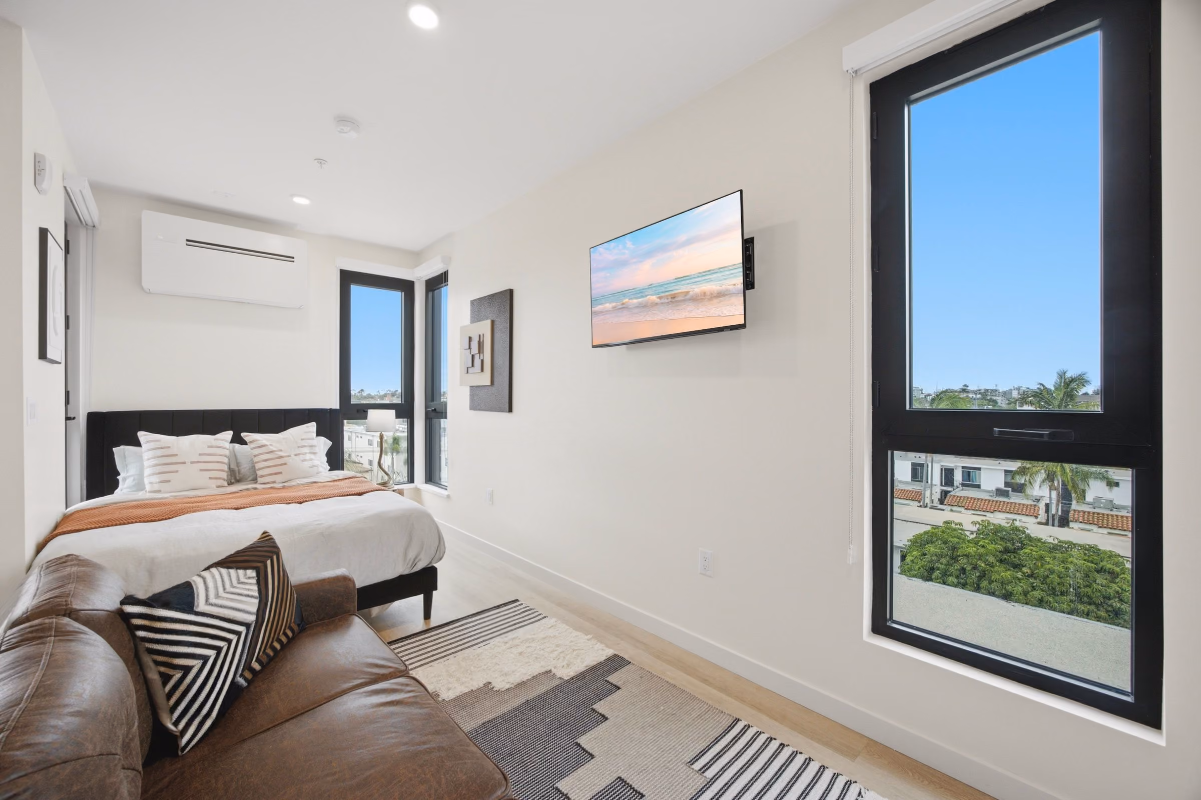 Bright bedroom with a bed, leather sofa with patterned pillow, wall-mounted TV, and large windows.