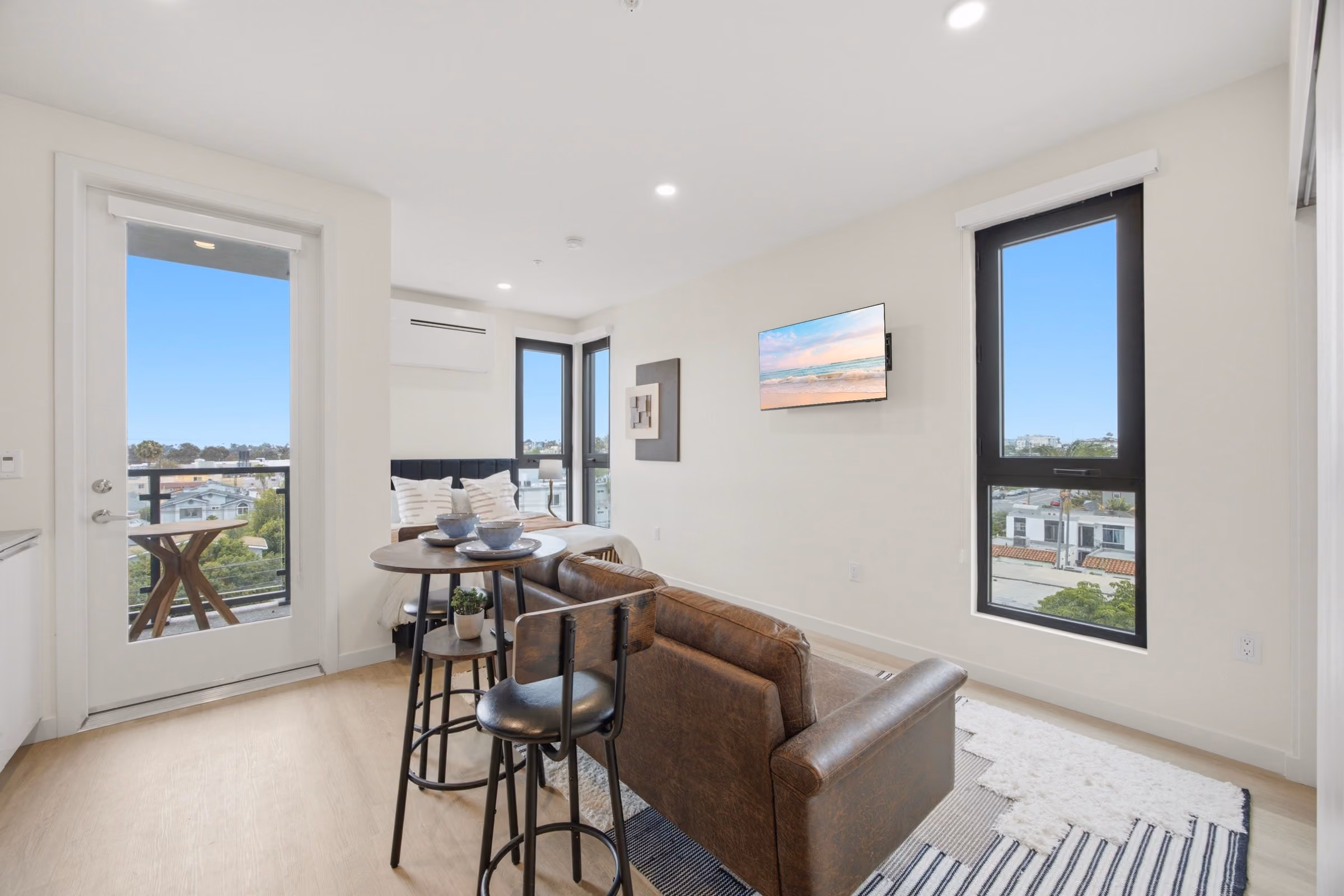 Modern studio apartment with bed, brown sofa, bar table, and windows with city views.