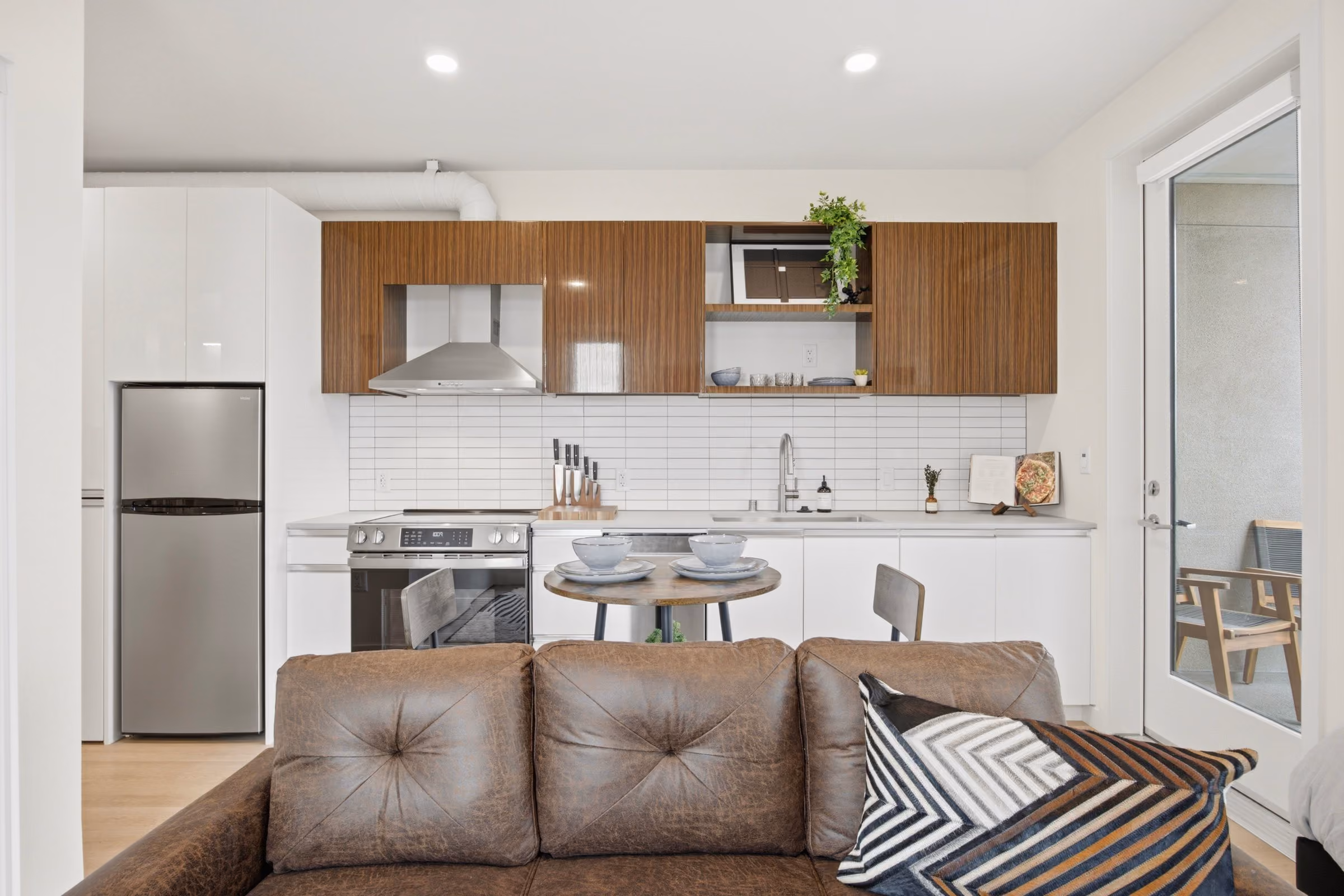 Modern kitchen with wood and white cabinets, stainless appliances, small dining table, and brown leather couch.