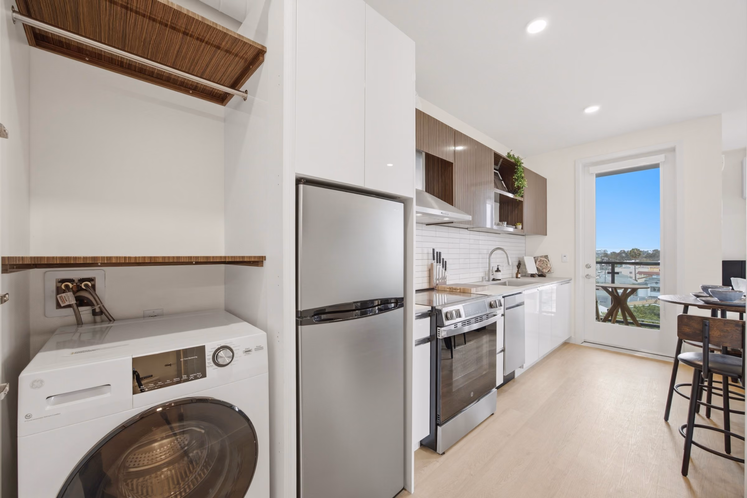 Modern kitchen with stainless steel appliances, washing machine, wooden cabinets, and door leading outside.