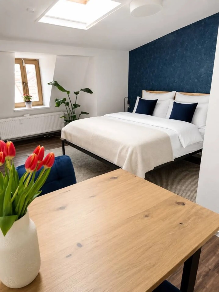 Helles Dachgeschoss-Schlafzimmer mit Doppelbett, blauer Akzentwand, Dachfenster und Holztisch mit roten Tulpen