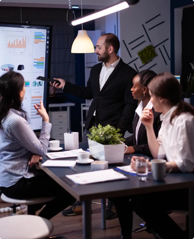Businessman in a suit presenting data charts on a digital screen to three colleagues seated around a table in an office meeting.