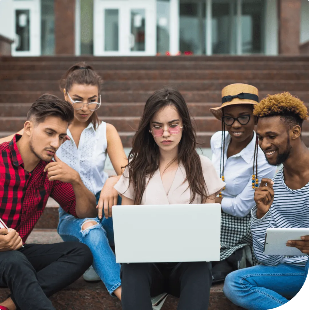 Group of five diverse young adults sitting on outdoor steps, collaborating around a laptop and tablet.