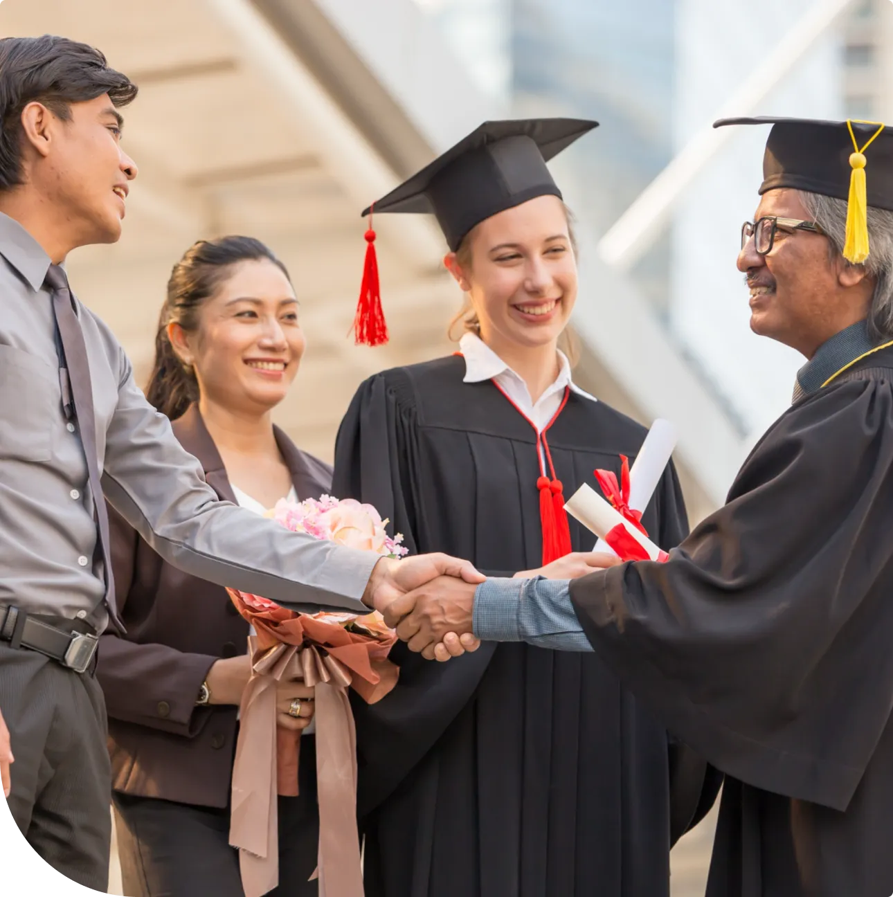 Smiling young female graduate in cap and gown receiving diploma and handshake from an older man in academic regalia, accompanied by two supporters.