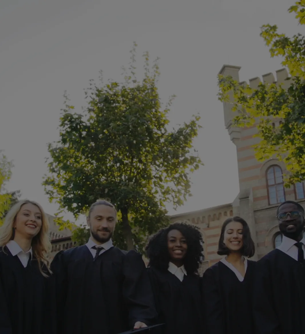 Group of diverse graduates in black gowns smiling outdoors in front of a historic building with trees.