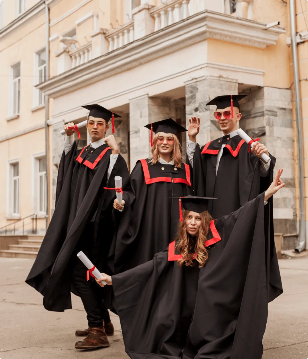 Four diverse graduates in black caps and gowns with red accents celebrating outdoors in front of a building, holding diplomas and smiling.