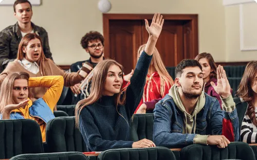 Students sitting in a classroom with two students raising their hands to answer or ask a question.