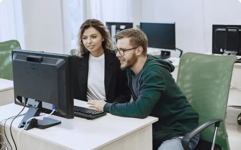 A man and a woman sit together at a desk looking at a computer monitor in an office setting.