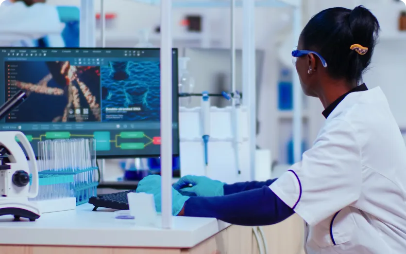 Scientist wearing gloves and protective glasses working at a lab computer displaying DNA and molecular structures.