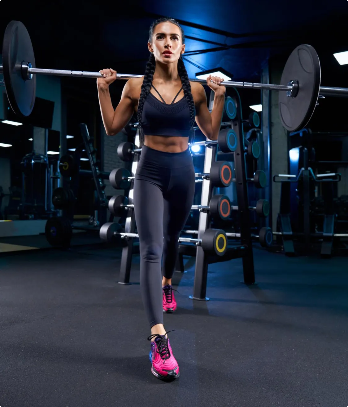 Woman doing barbell lunges in a gym, wearing black workout clothes and pink shoes.