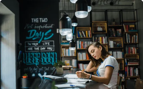 Young woman studying with a pencil and open books in a cozy library with hanging lights and a chalkboard wall.