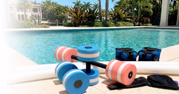 Aqua fitness equipment including water dumbbells, foam noodles, and resistance weights placed at the edge of a swimming pool for water workouts